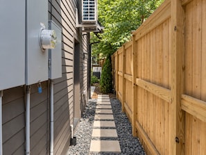 A narrow pathway with a wooden fence on one side and a building with outdoor electrical boxes on the other. - Maplewood Cottage • Heated Pool, Hot Tub, Gym, Grill, Fire Pit (Nashville)