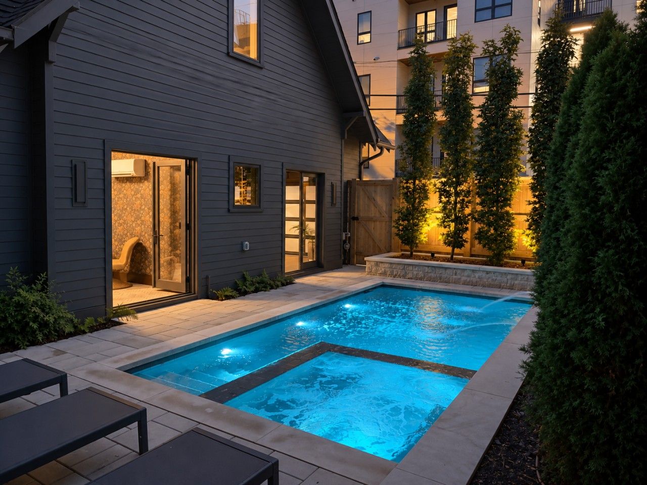 A swimming pool with a tiled deck, surrounded by a wooden fence and a building with a dark exterior.