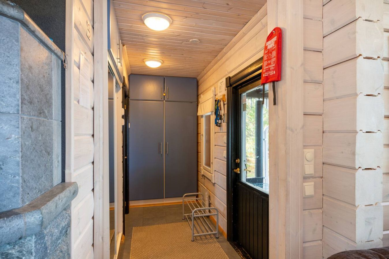 Un couloir étroit, avec une porte au fond, une main courante en métal et un panneau rouge sur le mur.