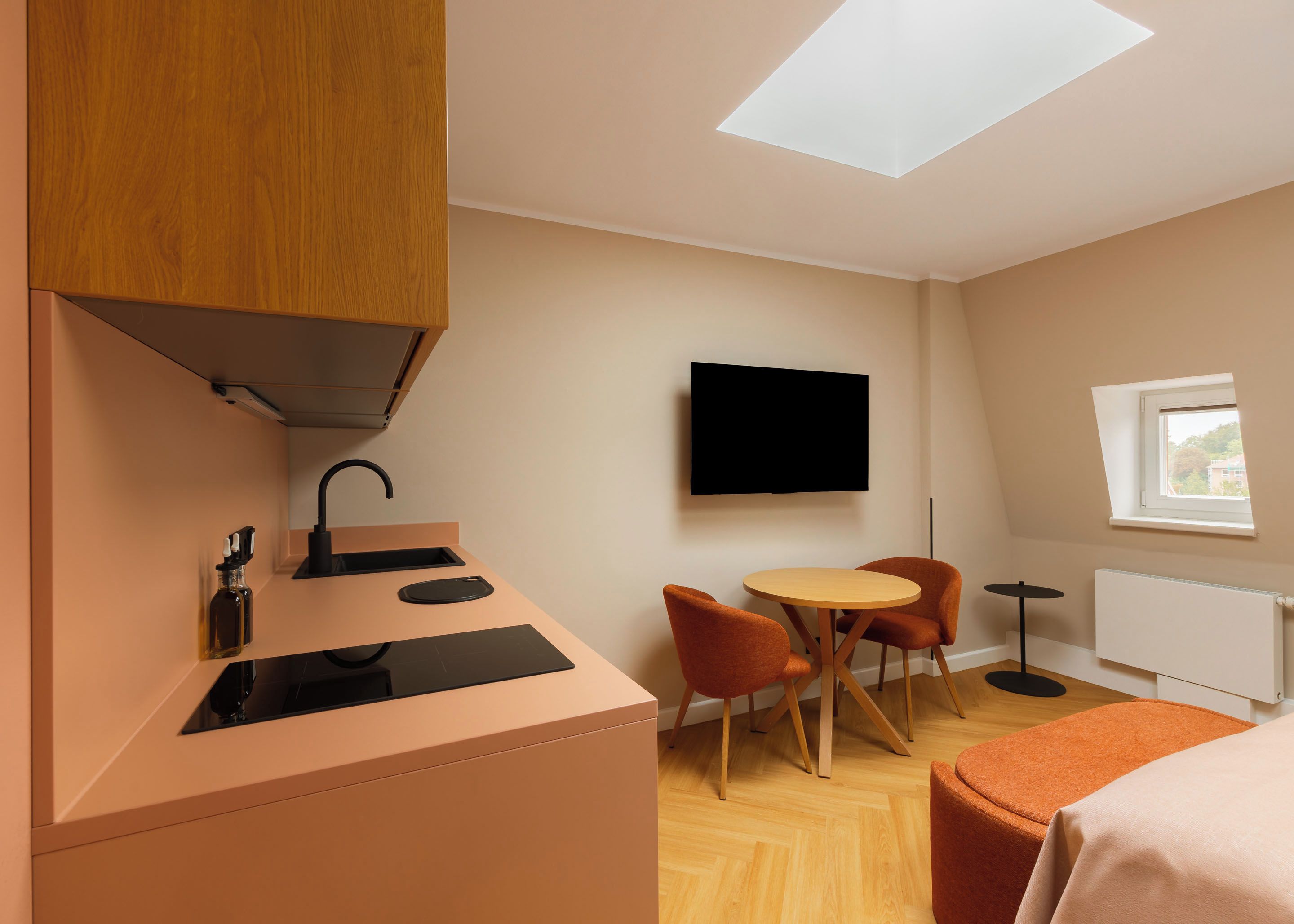 Eine kompakte Küchenzeile mit modernem Design, ein an der Wand montierter Flachbildfernseher, eine kleine Essecke mit zwei Stühlen und ein Fenster mit Tageslicht.