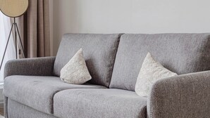 Un sofà gris amb dos coixins decoratius, una làmpada de peu i una finestra amb cortines.