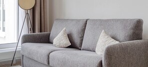 Ein graues Sofa mit zwei Zierkissen, einer Stehlampe und einem Fenster mit Vorhängen.