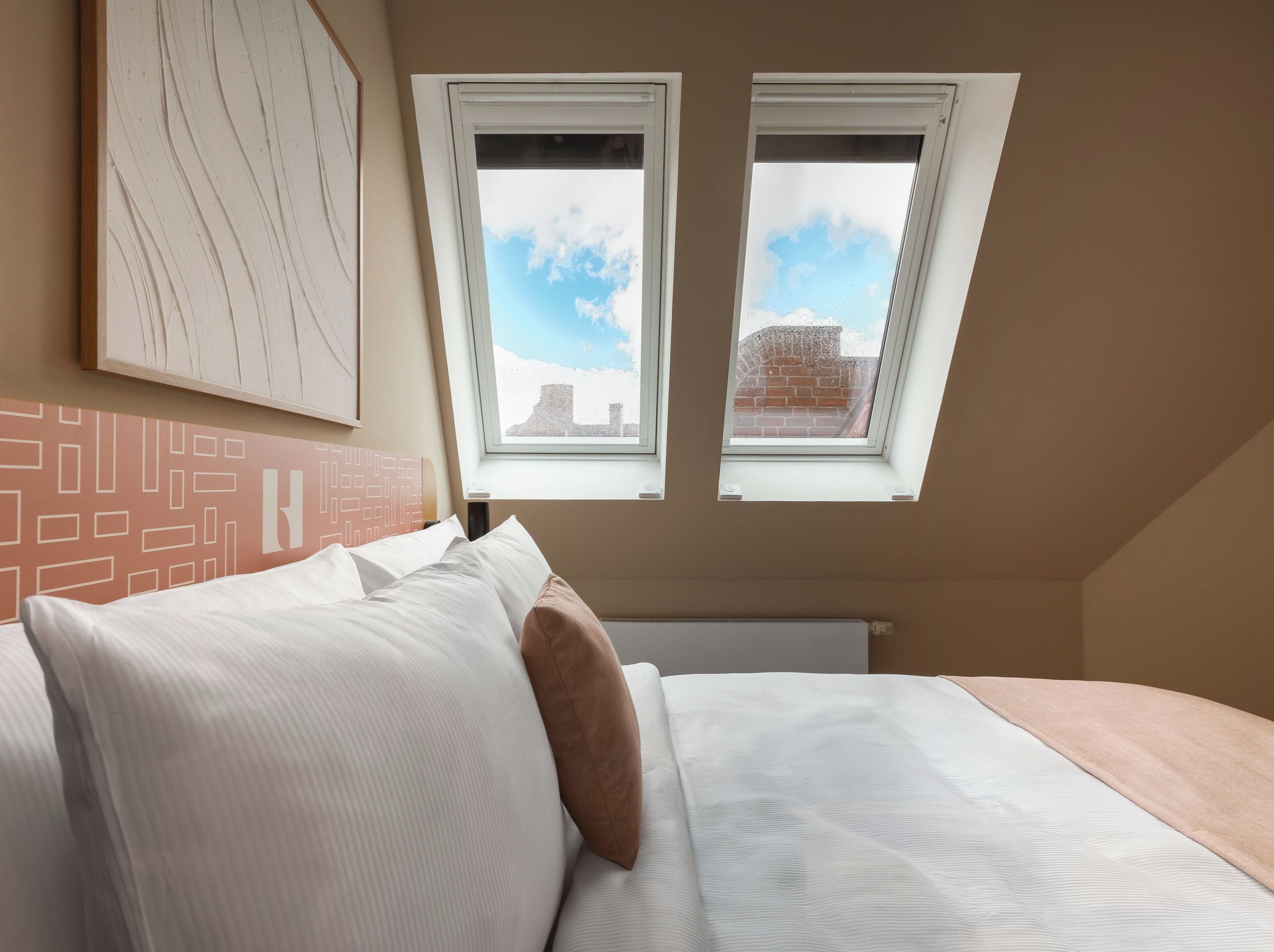 Ein Schlafzimmer mit einem Bett, Kissen, einem Kopfteil mit geometrischem Muster und zwei Fenstern mit Blick auf eine Backsteinmauer und den Himmel.