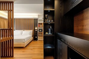 Een moderne hotelkamer met een houten spijlenwand, een bed met witte lakens, een klein nachtkastje, een plank met boeken en een koffiezetapparaat, en een donker houten kast.