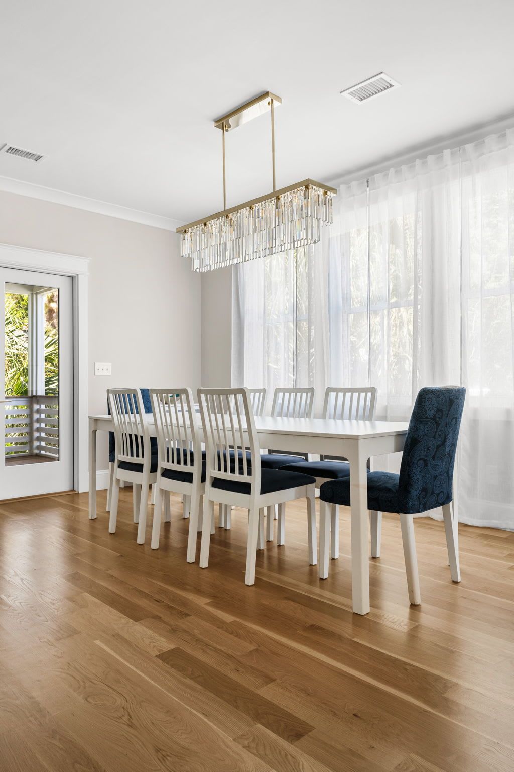 A dining area with a white table and chairs, a chandelier, and a window with sheer curtains.