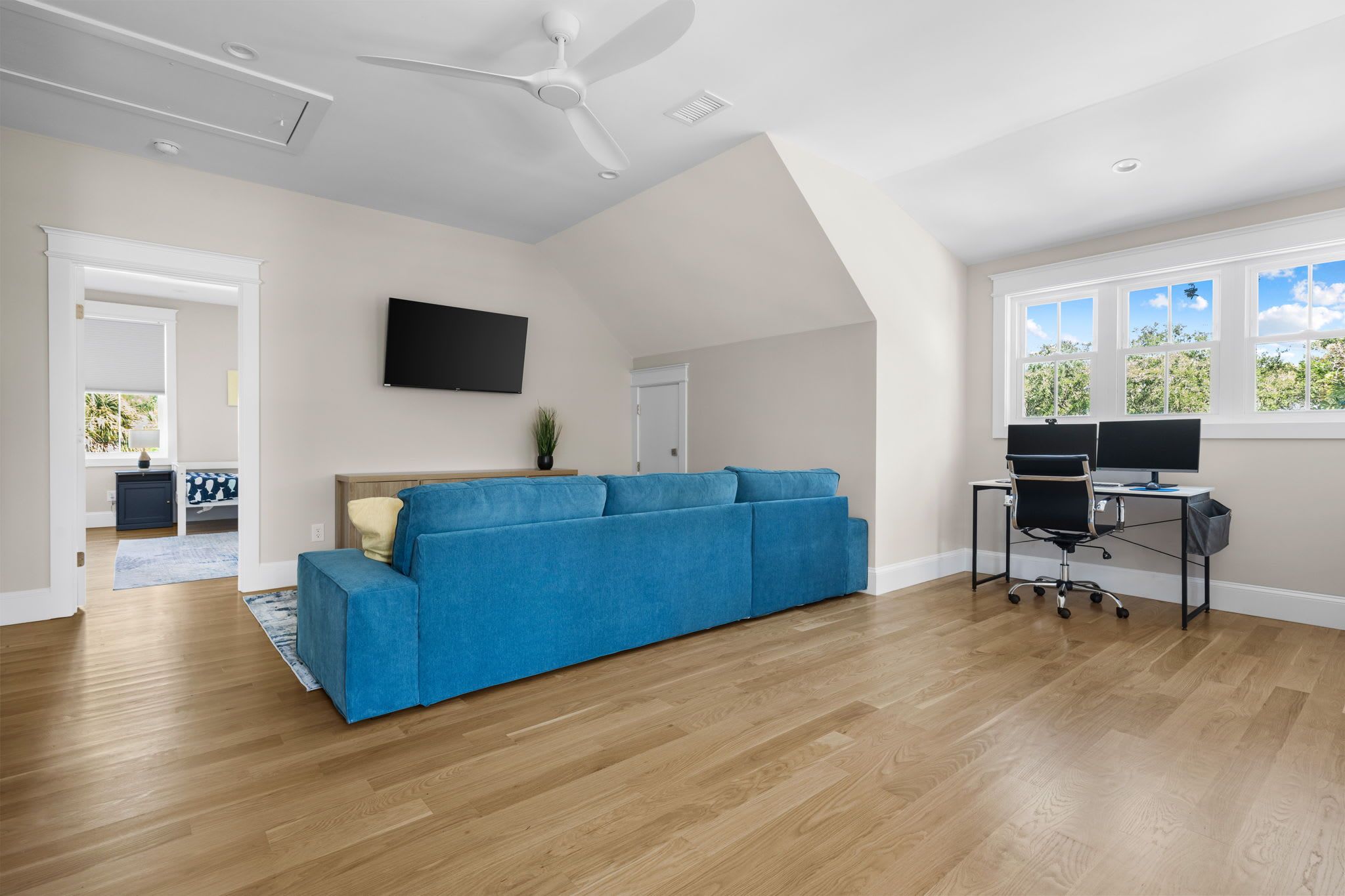 A spacious living room with a blue sofa, a ceiling fan, and a large window with a view of greenery.