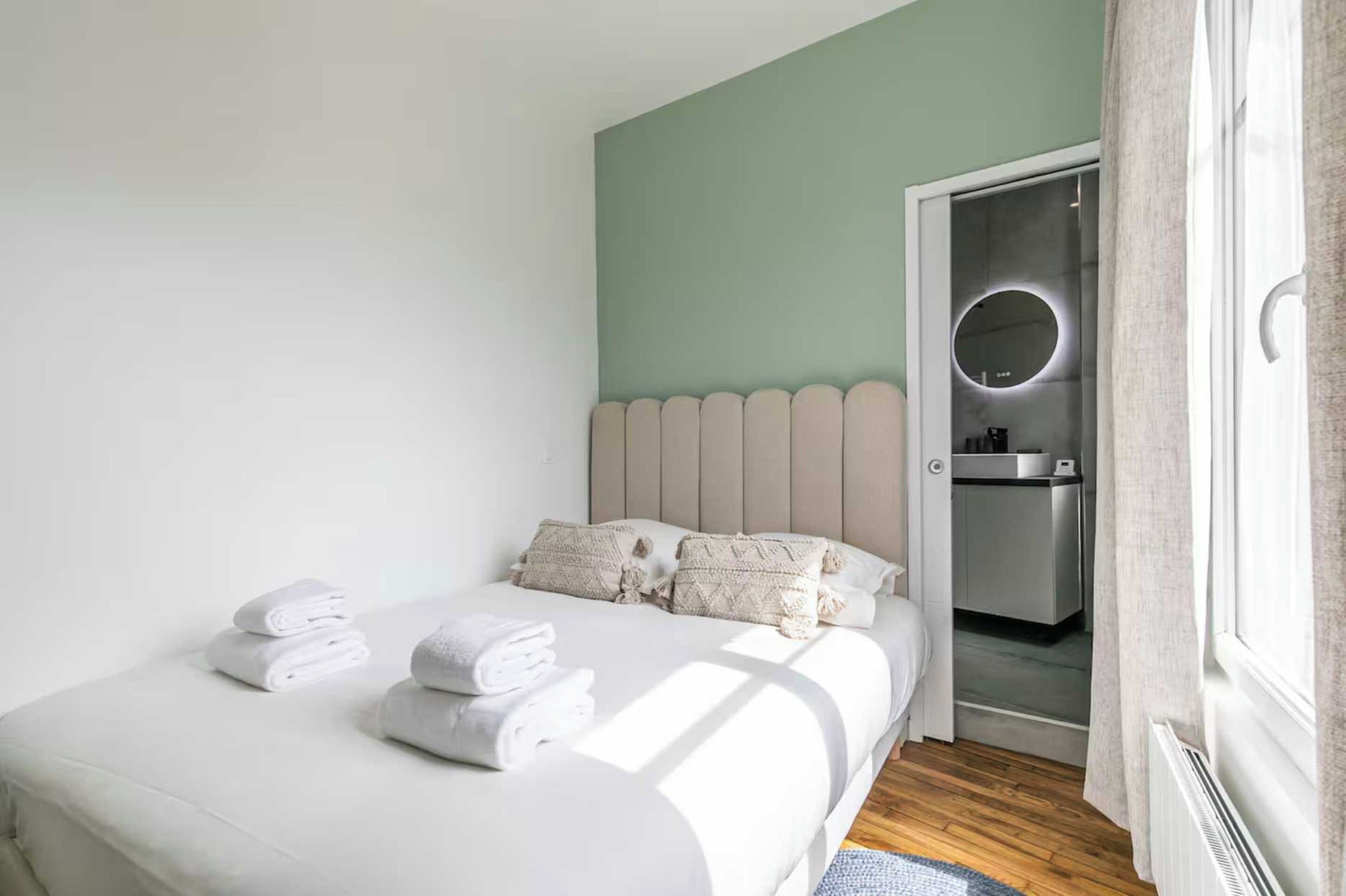 Un dormitorio con cama, almohadas y toallas dobladas. Hay una puerta que da a un baño con espejo y lavamanos.