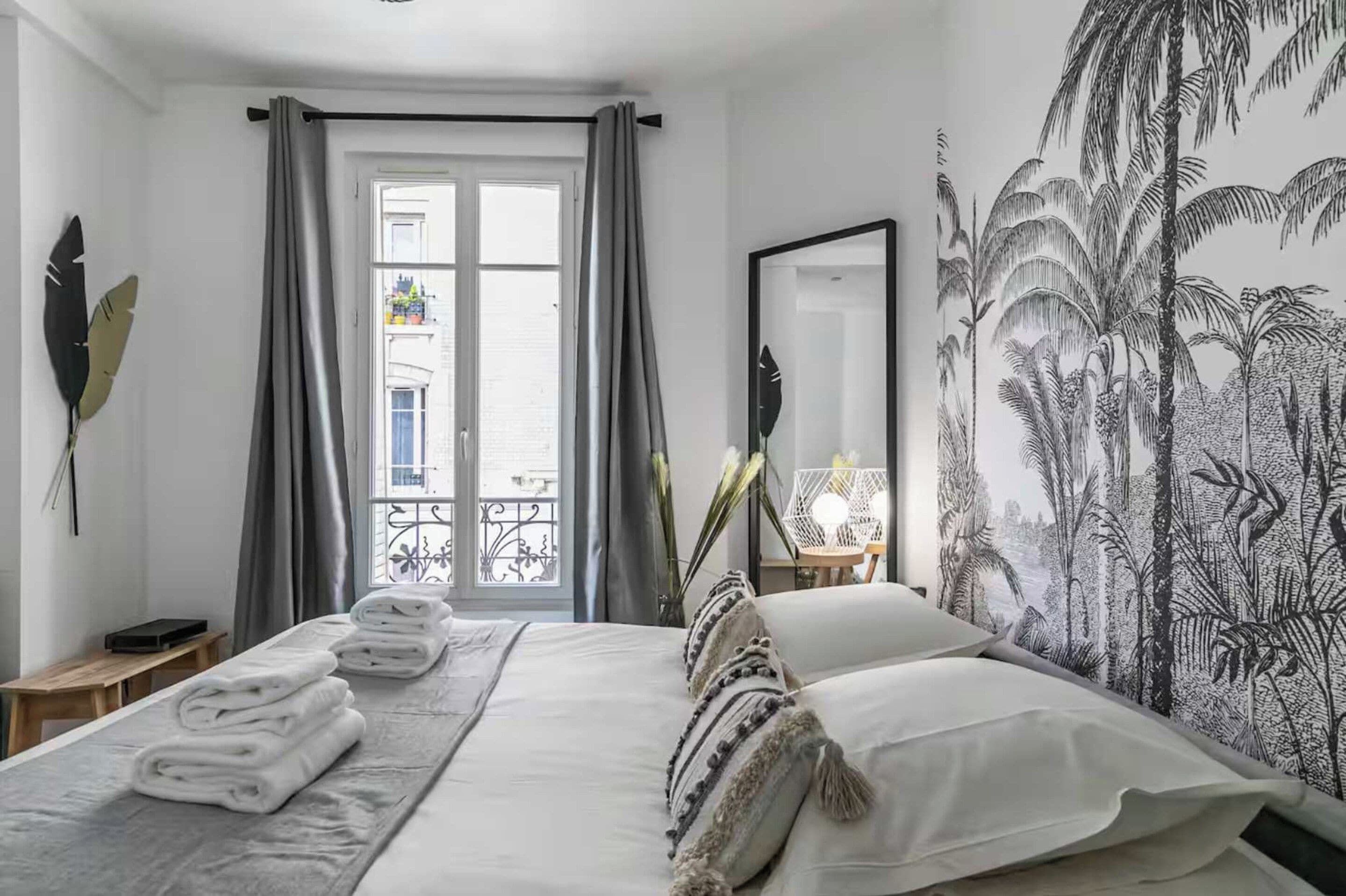 A hotel room with a bed, a window with curtains, a mirror, and a wall with a plant pattern.
