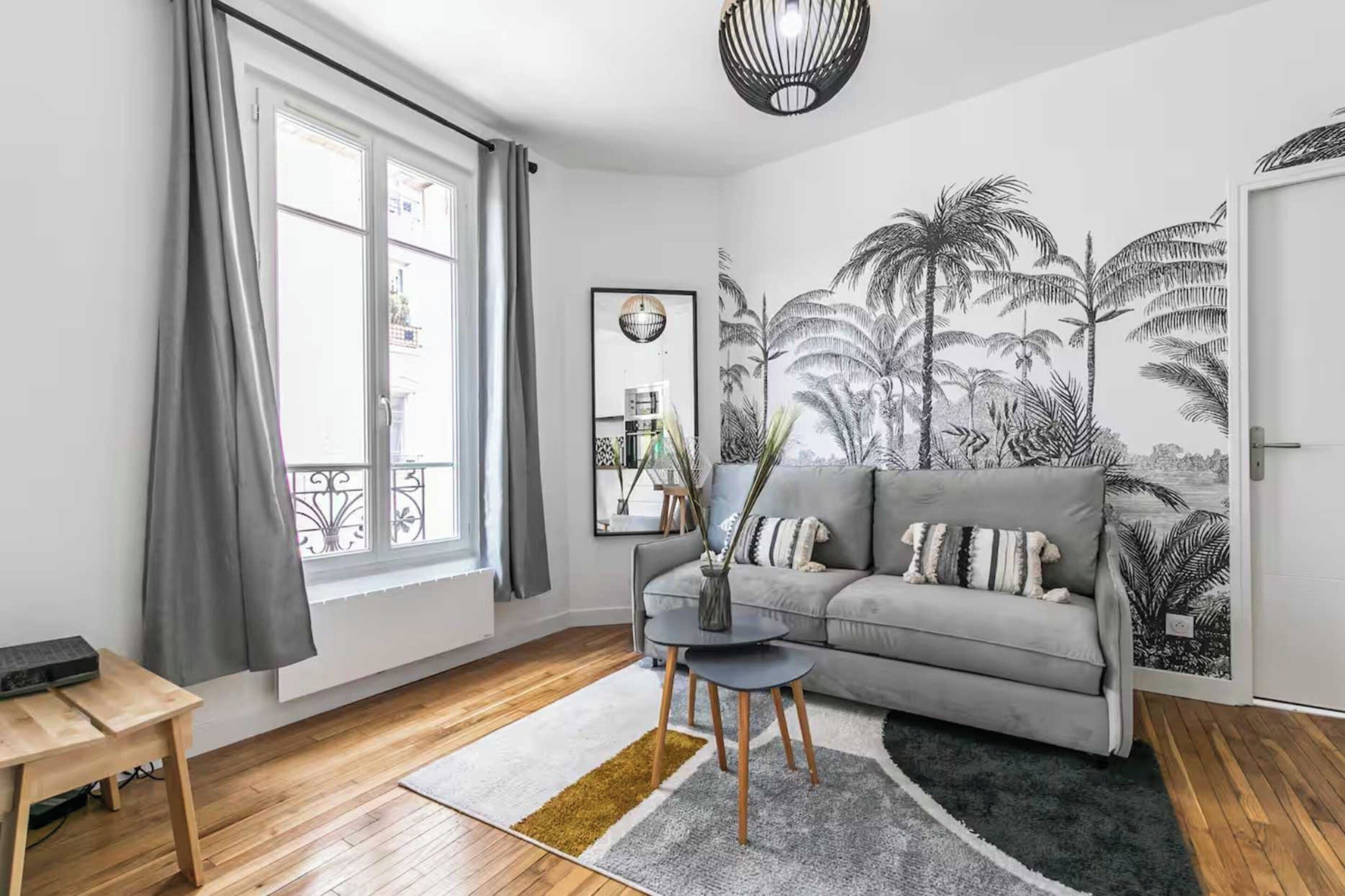 Een moderne woonkamer met een grijze bank, een klein rond tafeltje en een groot raam met gordijnen.
