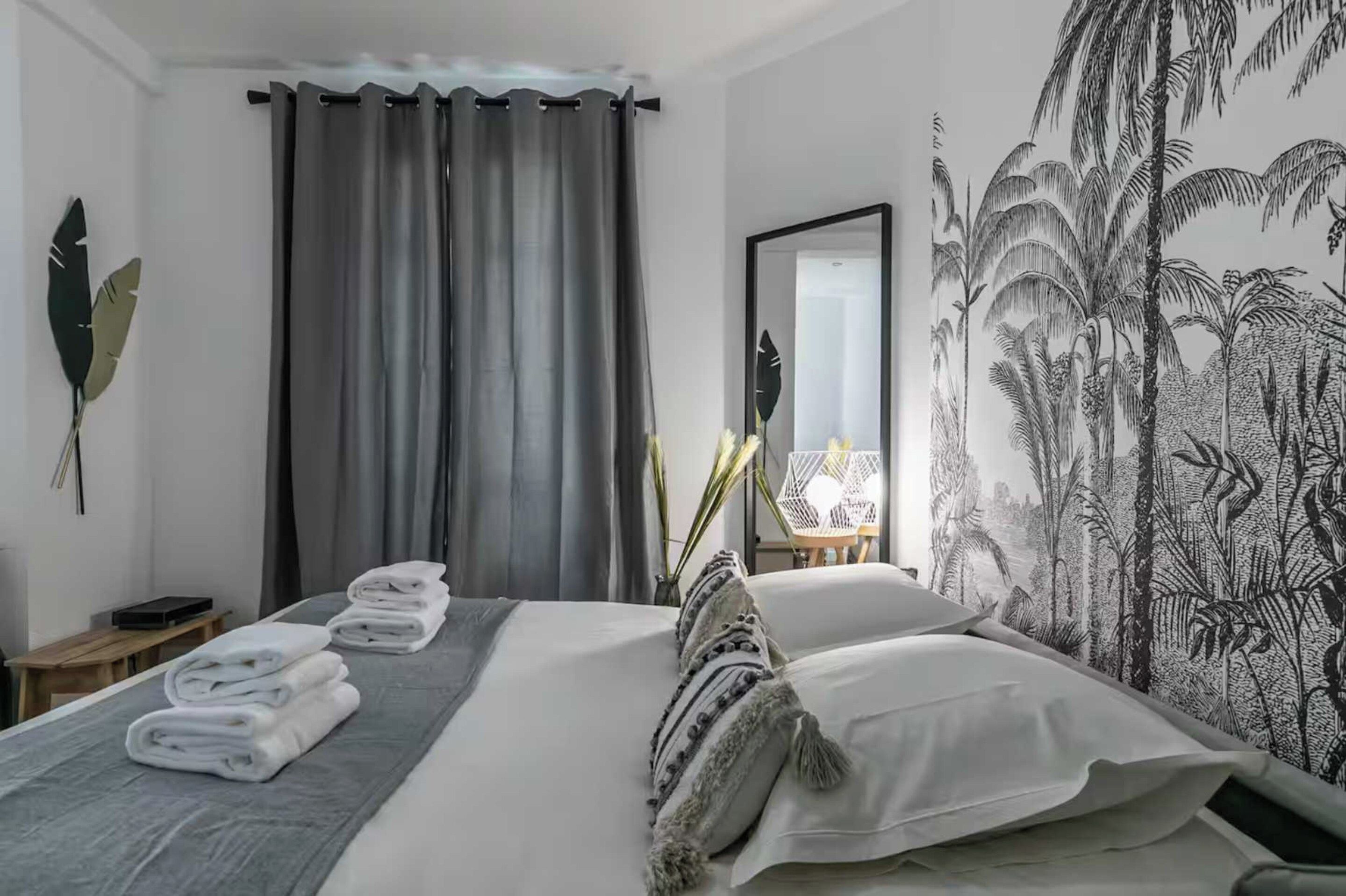 A hotel room with a bed, a mirror, a wall with a nature-themed mural, and a window with grey curtains.