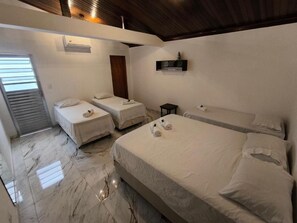 A hotel room with a large bed, two smaller beds, a bedside table, a wall-mounted shelf, and a window with blinds. - Casa no Centro Histórico de Cananéia – Perto de Tudo e da Beira-Mar (Cananéia)