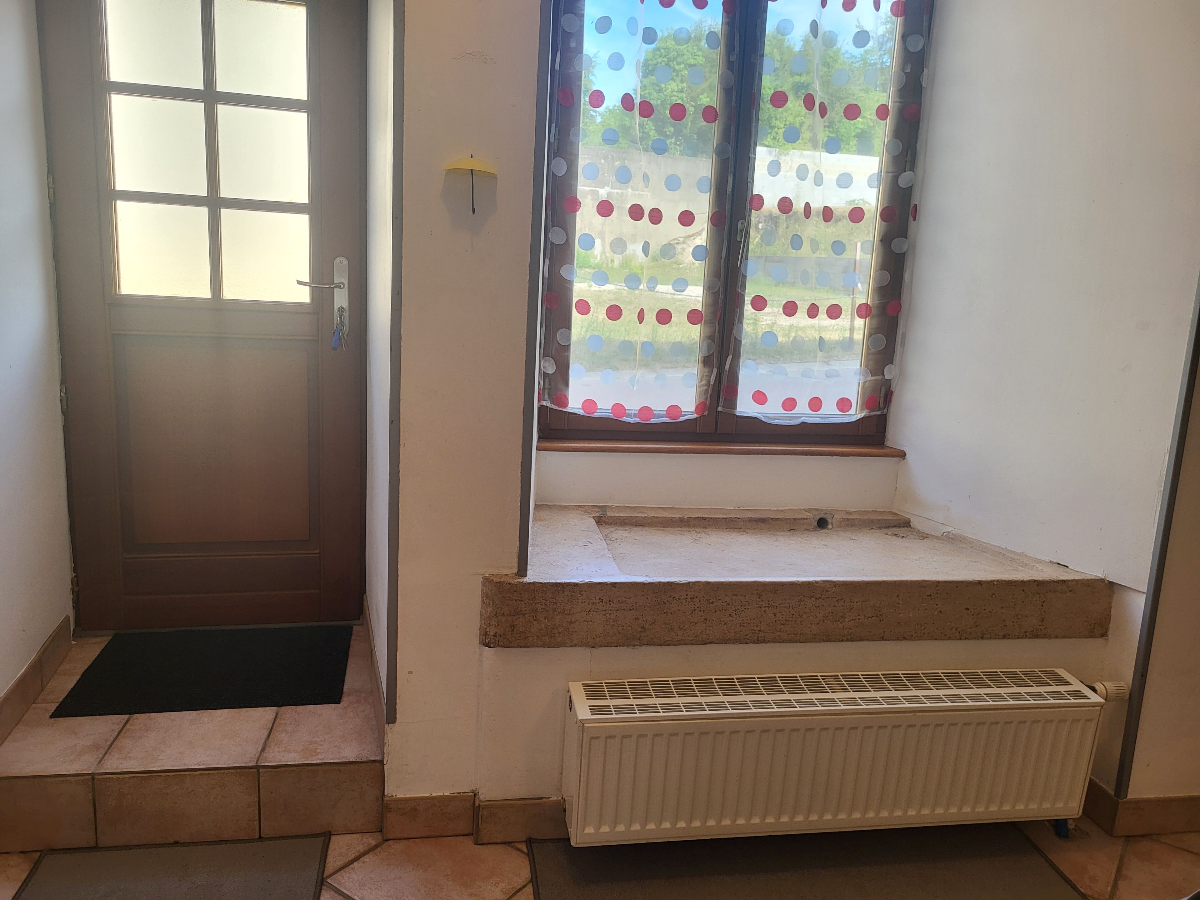 Une pièce avec une porte, une fenêtre ornée d’un rideau à pois, un radiateur et un petit marchepied menant à un palier.