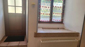 Une pièce avec une porte, une fenêtre ornée d’un rideau à pois, un radiateur et un petit marchepied menant à un palier.