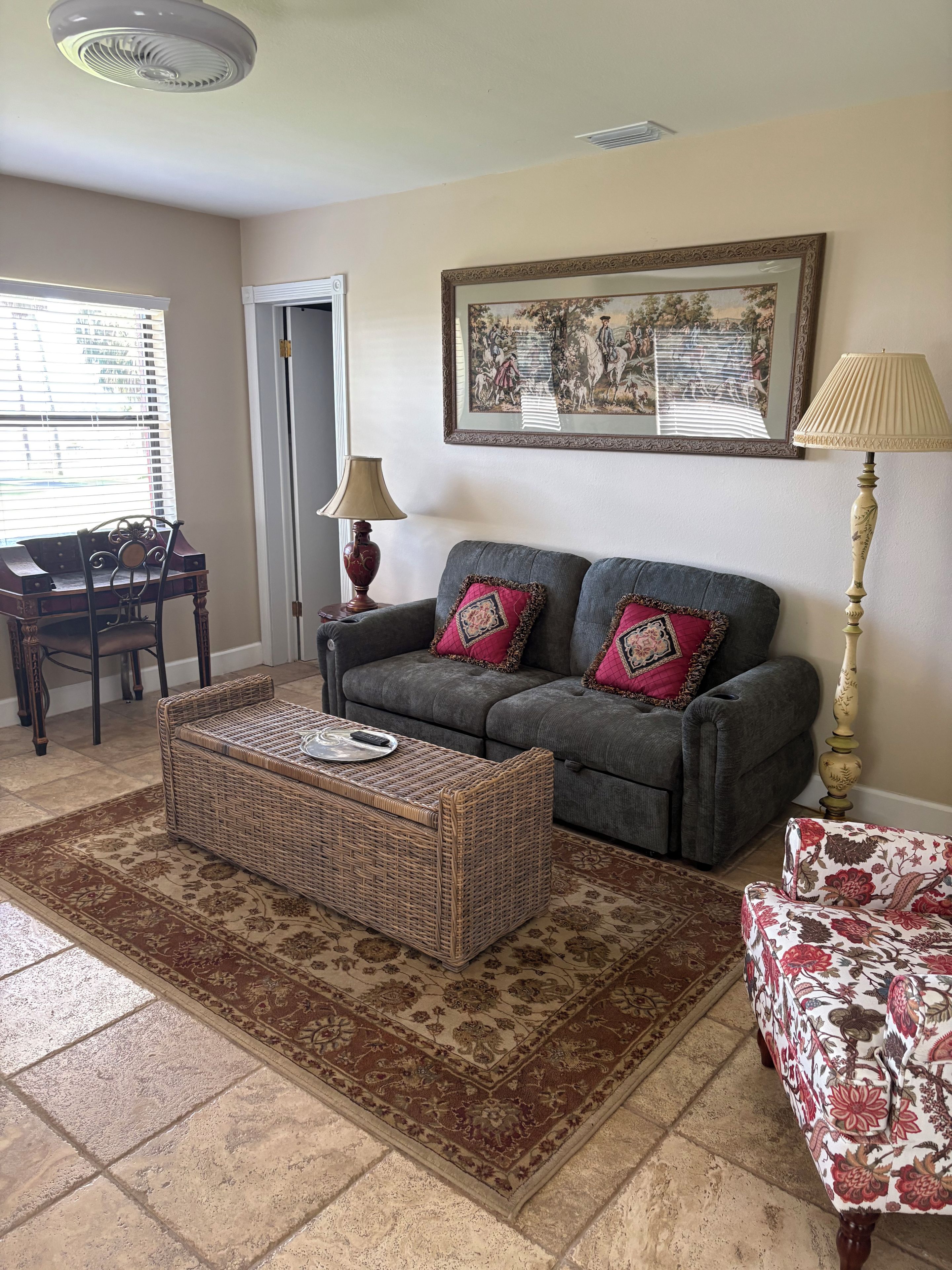 Un salon avec un canapé gris, deux coussins décoratifs, une table basse en osier, un tapis à motifs, une chaise en bois, une lampe et un tableau encadré accroché au mur.