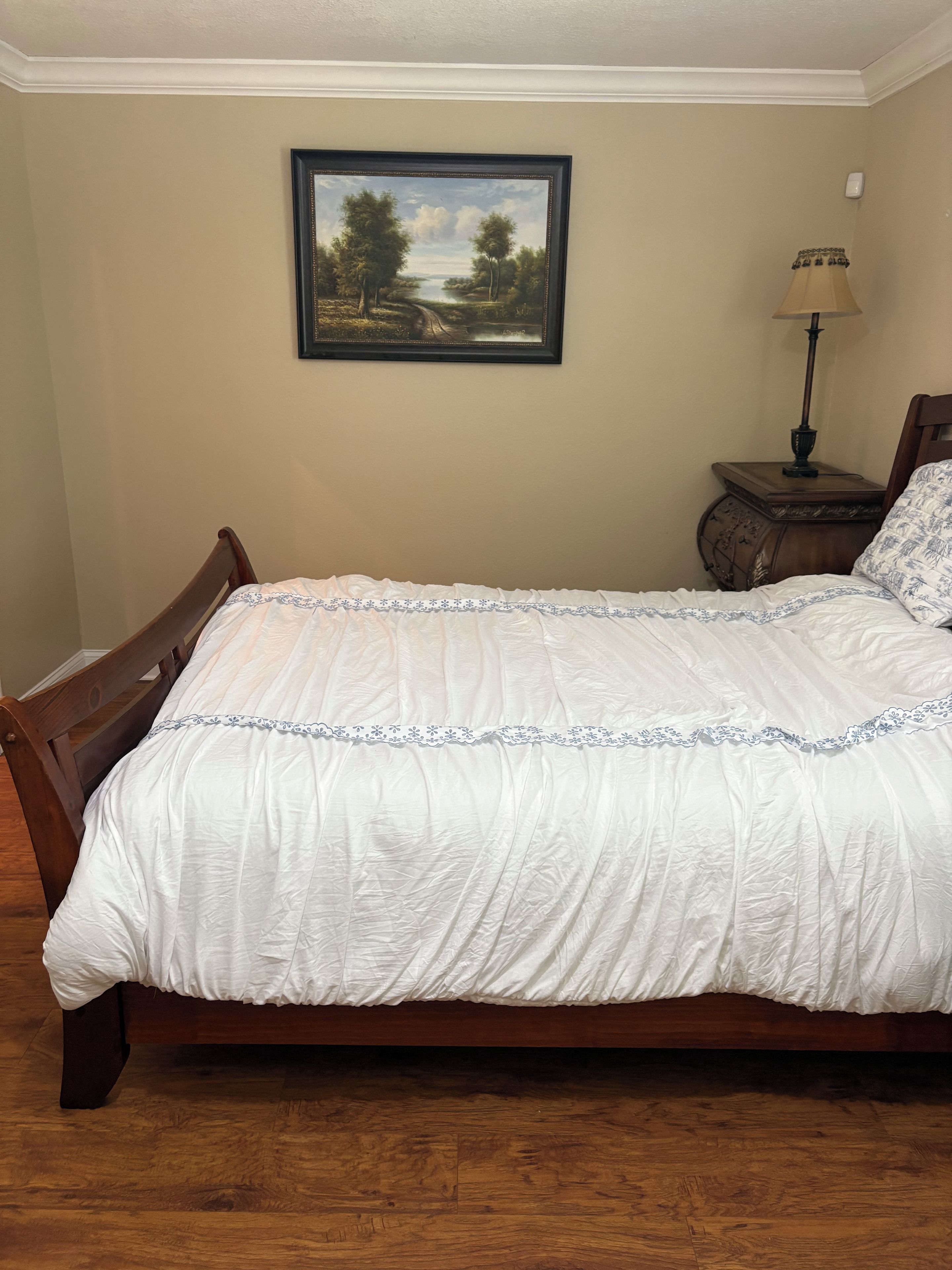 A bedroom with a wooden bed, a nightstand with a lamp, and a framed landscape painting on the wall.