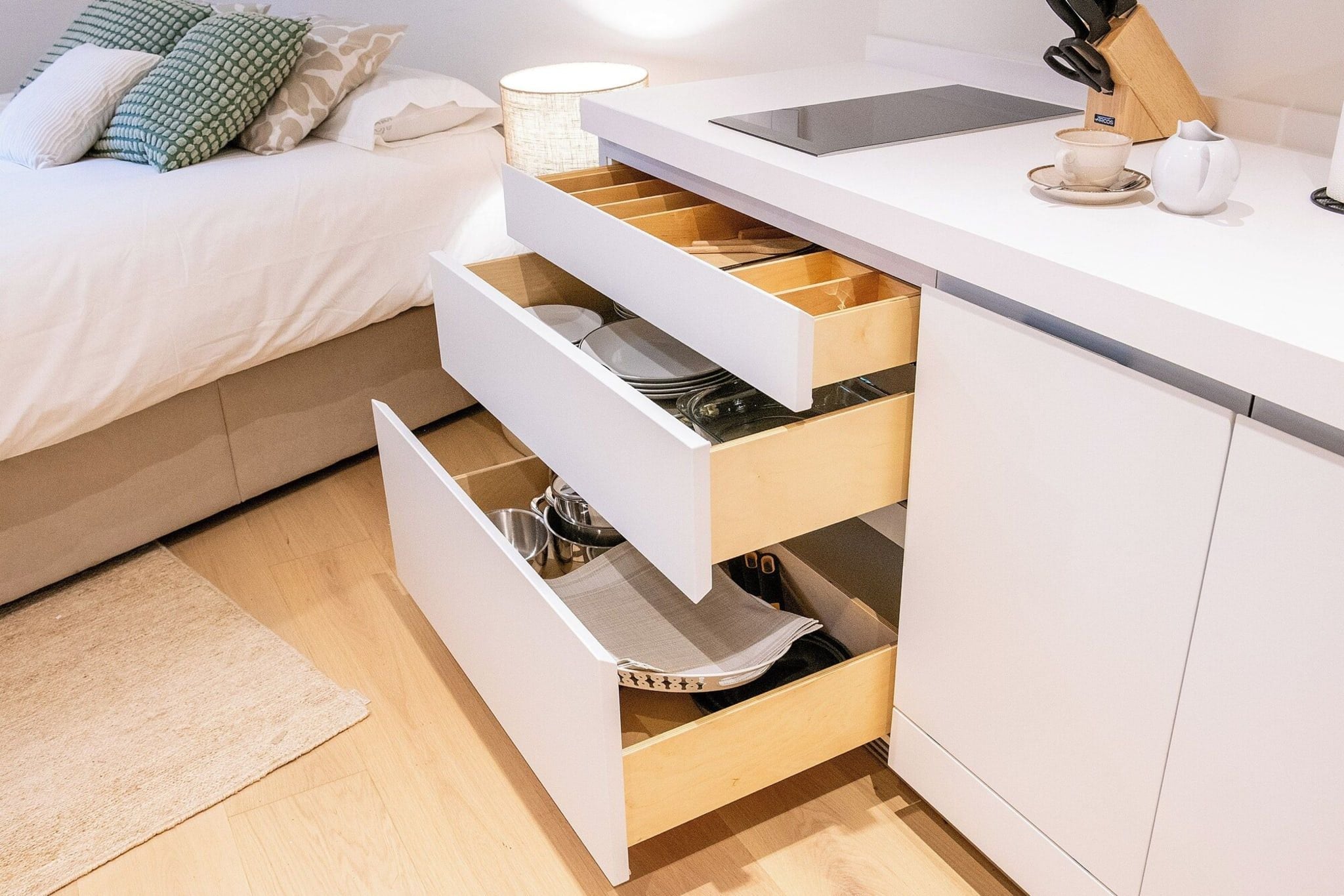 Eine moderne Küche mit weißen Schränken, einer Arbeitsplatte und einem Bett im Hintergrund, ausgestattet mit weißer Bettdecke und gemusterten Kissen.