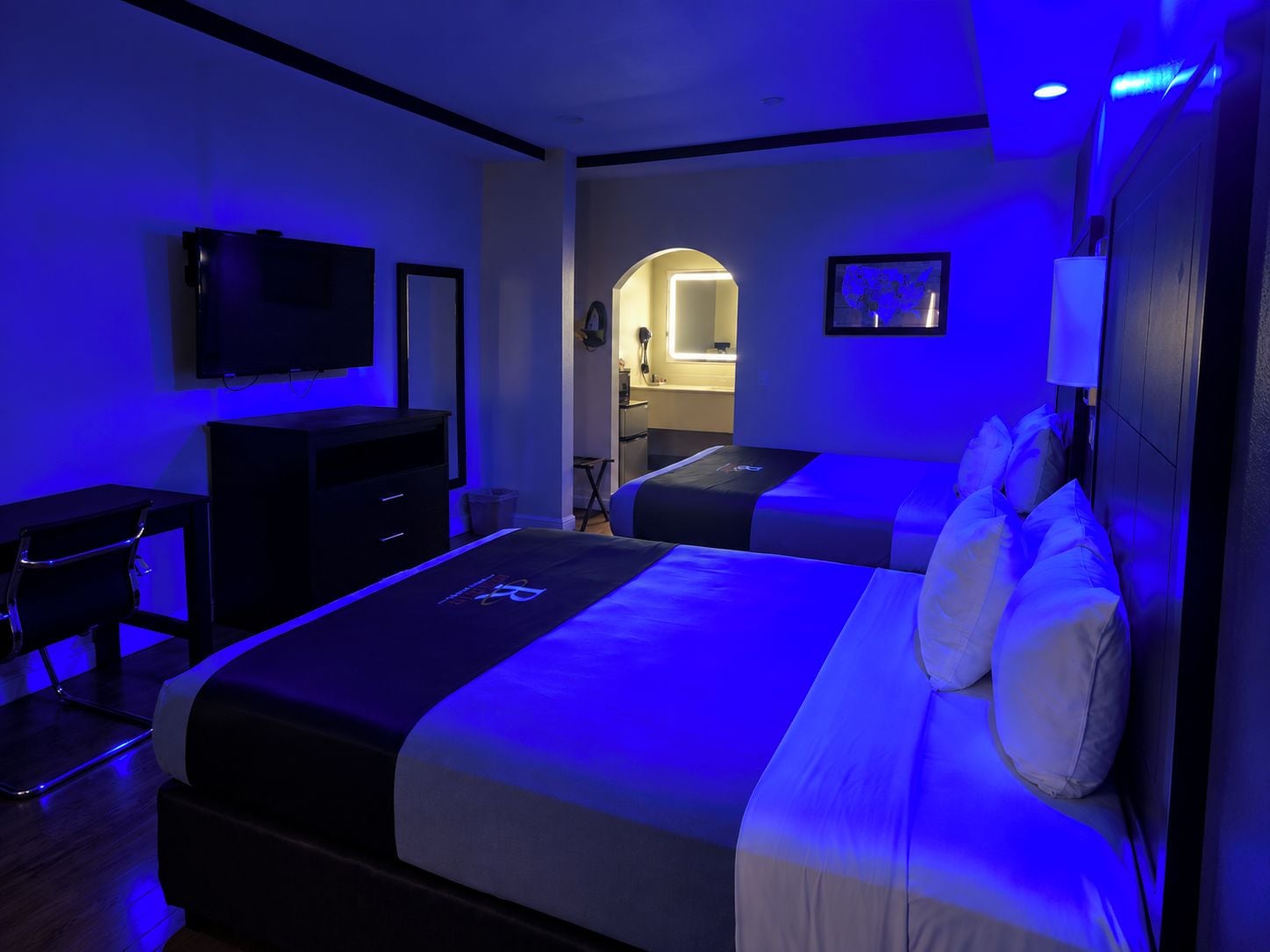 A hotel room with two beds, a desk, a TV, and a bathroom visible through an archway.