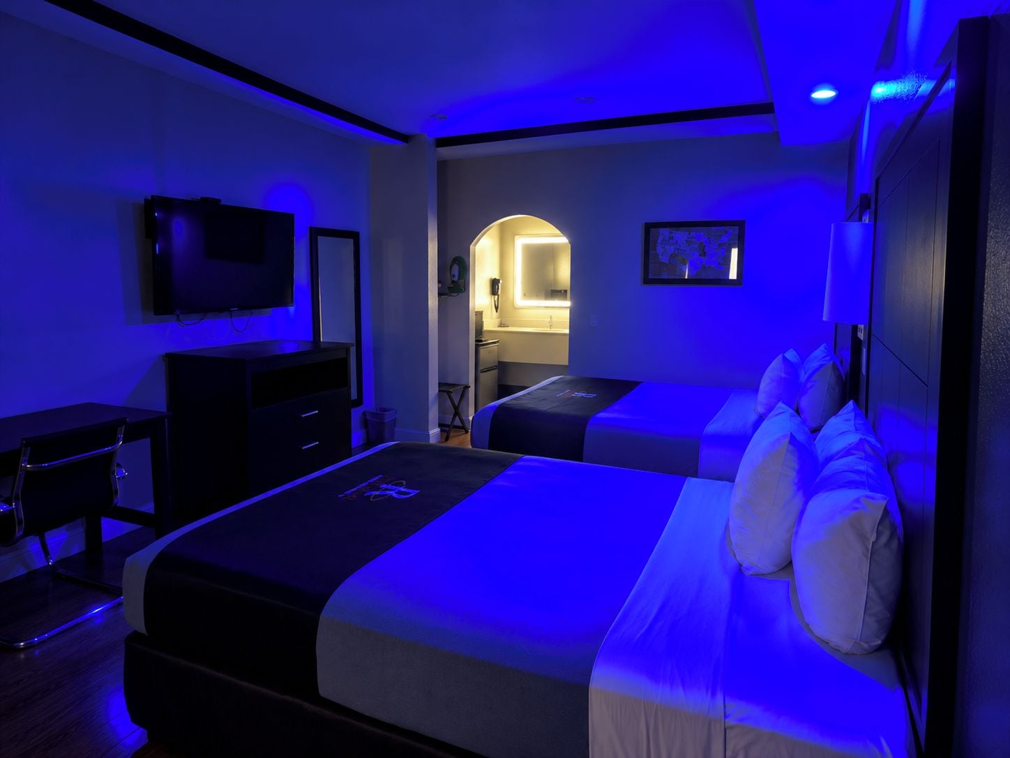 A hotel room with a bed, a desk, a chair, a television, and a bathroom visible through an archway.