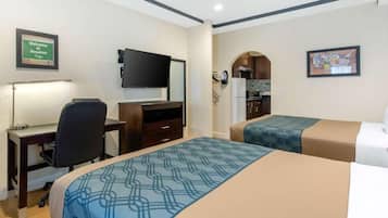 A hotel room with a desk, a flat-screen TV, a bed with patterned bedding, a kitchenette, and a framed welcome sign.