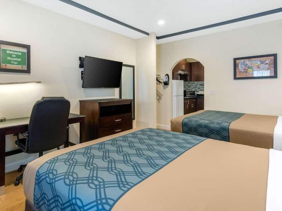 Habitación de hotel con escritorio, televisor de pantalla plana, cama con ropa de cama estampada, kitchenette y cartel de bienvenida enmarcado.
