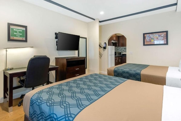A hotel room with a desk, a flat-screen TV, a bed with patterned bedding, a kitchenette, and a framed welcome sign.