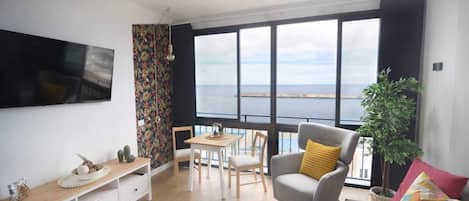 Een moderne woonkamer met een groot raam, een flatscreen televisie, een eethoek en een bank met kussens.