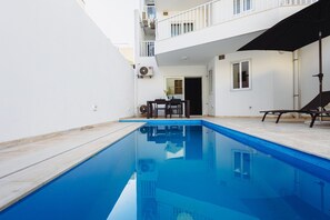 A swimming pool with a clear blue water surface, surrounded by a white building with balconies and a covered outdoor seating area. - Modern Maisonette with Pool in Gharghur (Ħal Għargħur)