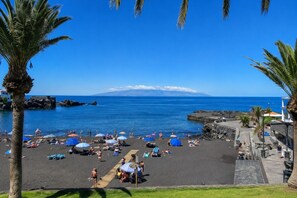 Een strand met mensen die zonnebaden, parasols en een helder uitzicht op de oceaan en een berg in de verte.