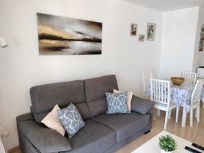 Een moderne woonkamer met een grijze bank, gekleurde kussens, een schilderij en een eethoek met witte stoelen en een tafel.