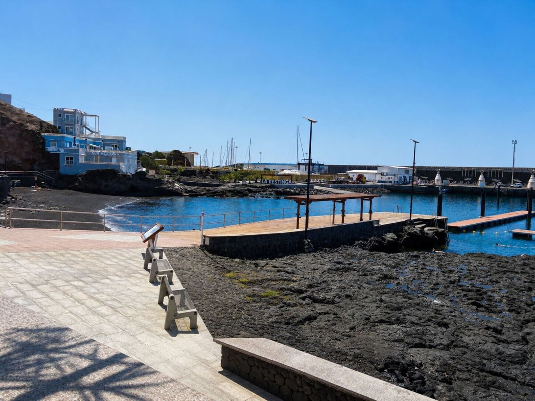 Uma marina costeira com céu claro, um litoral rochoso e um edifício com a inscrição 'LA RESTINGA'.