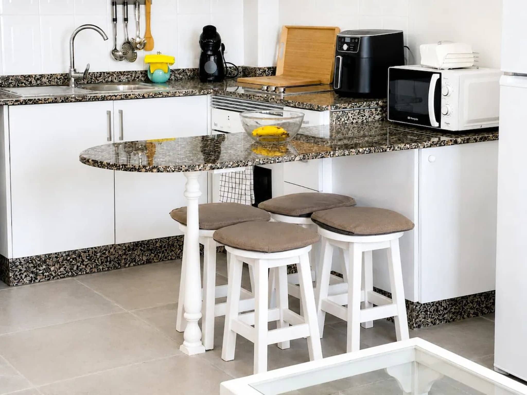 A kitchen with a granite countertop, white cabinets, a coffee maker, a toaster, and a microwave. There are three stools with cushions.