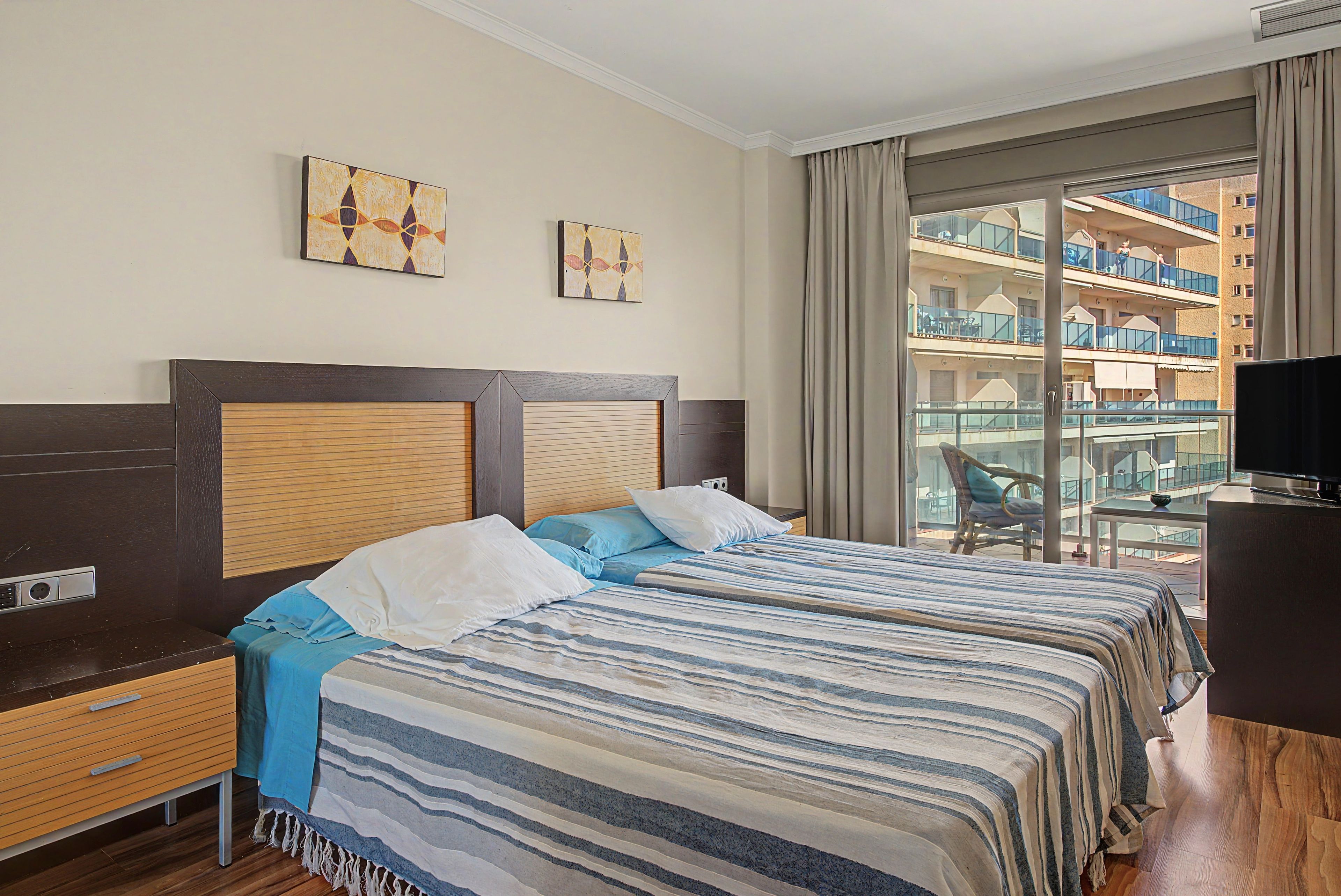 A hotel room with two beds, a wooden headboard, a TV, and a balcony with a view of the building.