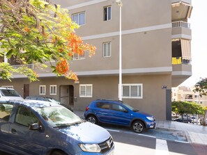 A multi-story residential building with balconies, parked cars, and a tree with orange flowers. - CHL Casa Arthur, ático en El Médano cerca de la pl (El Médano)