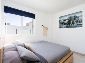 Ein Schlafzimmer mit einem Bett, einem Fenster mit blauer Jalousie, einer Wanddekoration aus Makramee und einem Bilderrahmen mit Elefantenmotiven.