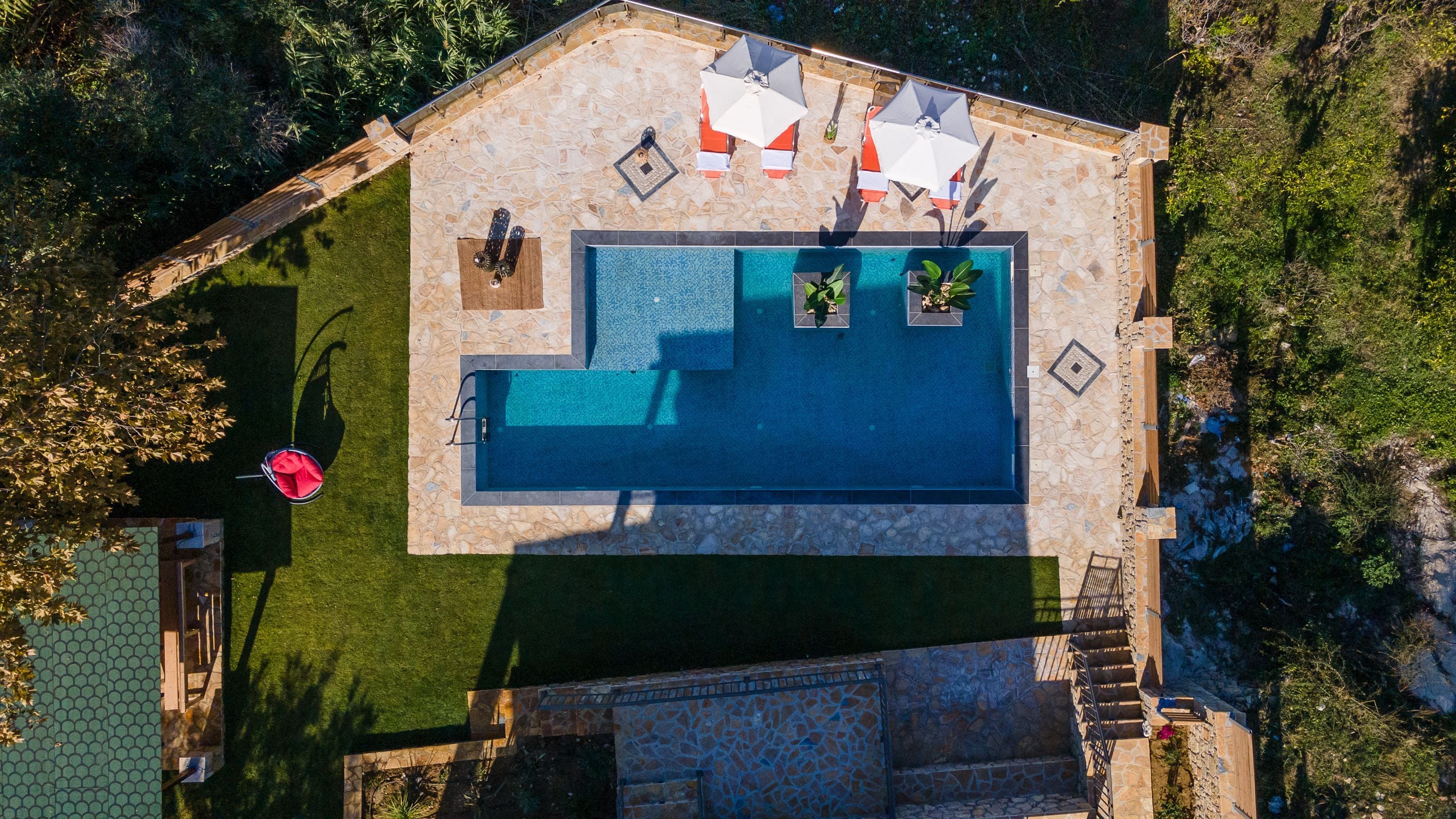 Vista aérea de una piscina con plataforma de piedra, rodeada de un césped cuidado y algunas macetas con plantas.