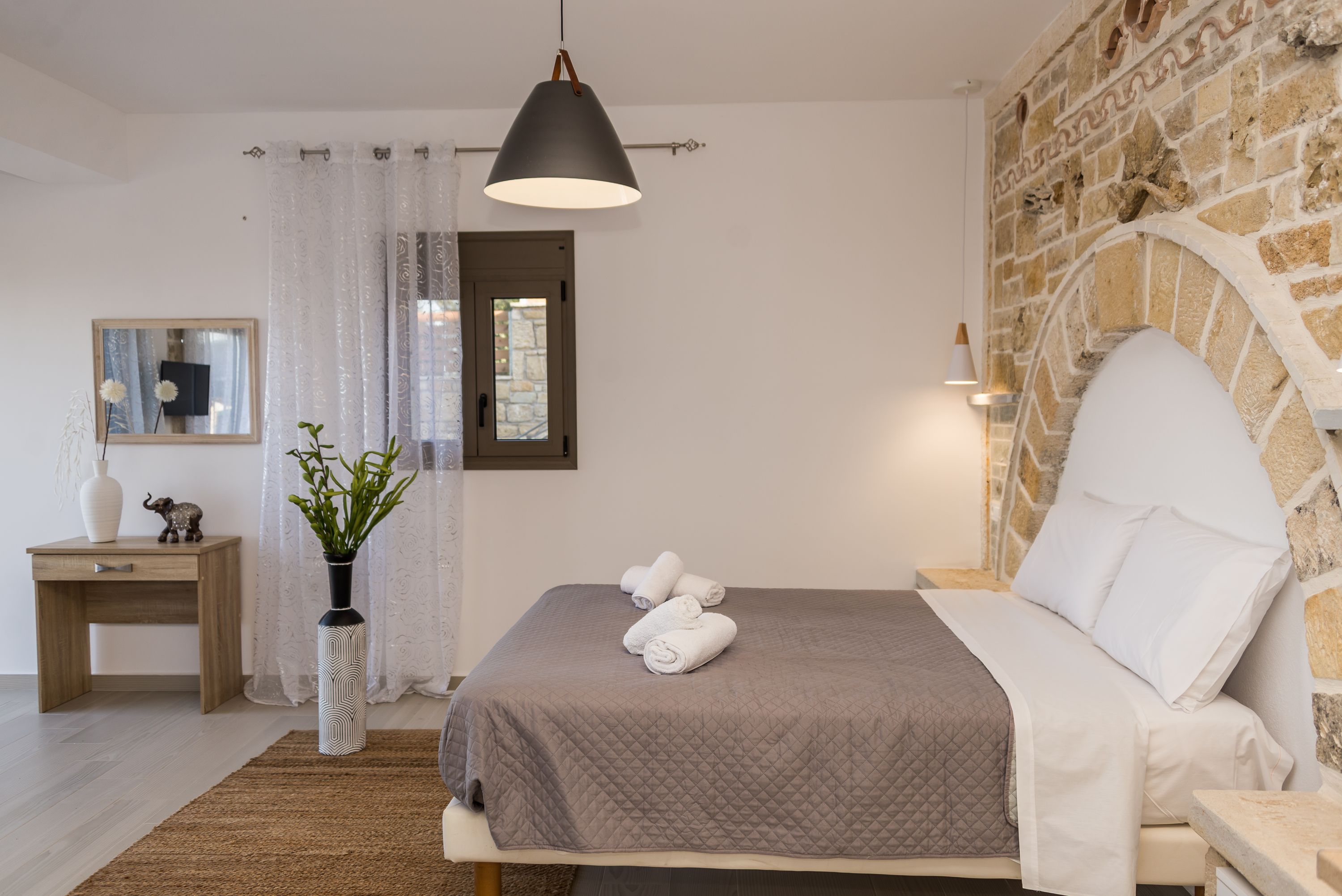 Un dormitorio con cama, mesita de noche, espejo, jarra con plantas y pared de piedra.
