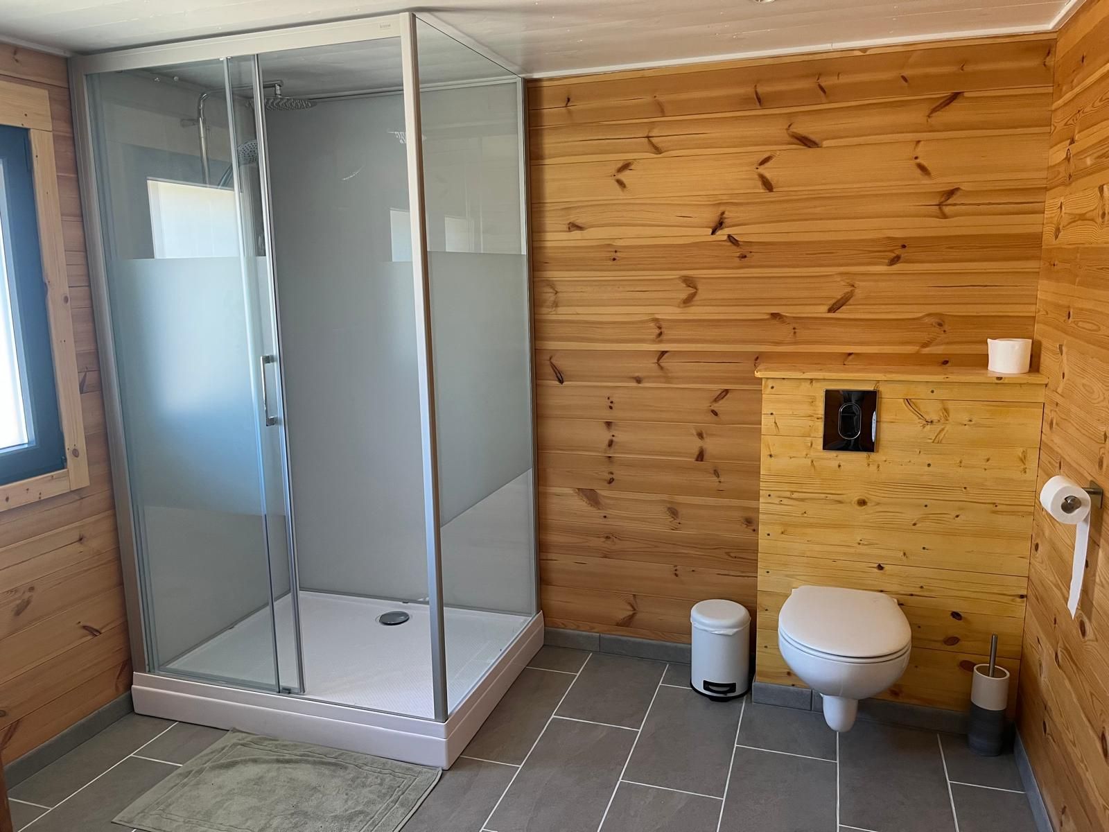 A modern bathroom with a glass-enclosed shower, wooden panel walls, a toilet, and a trash bin.