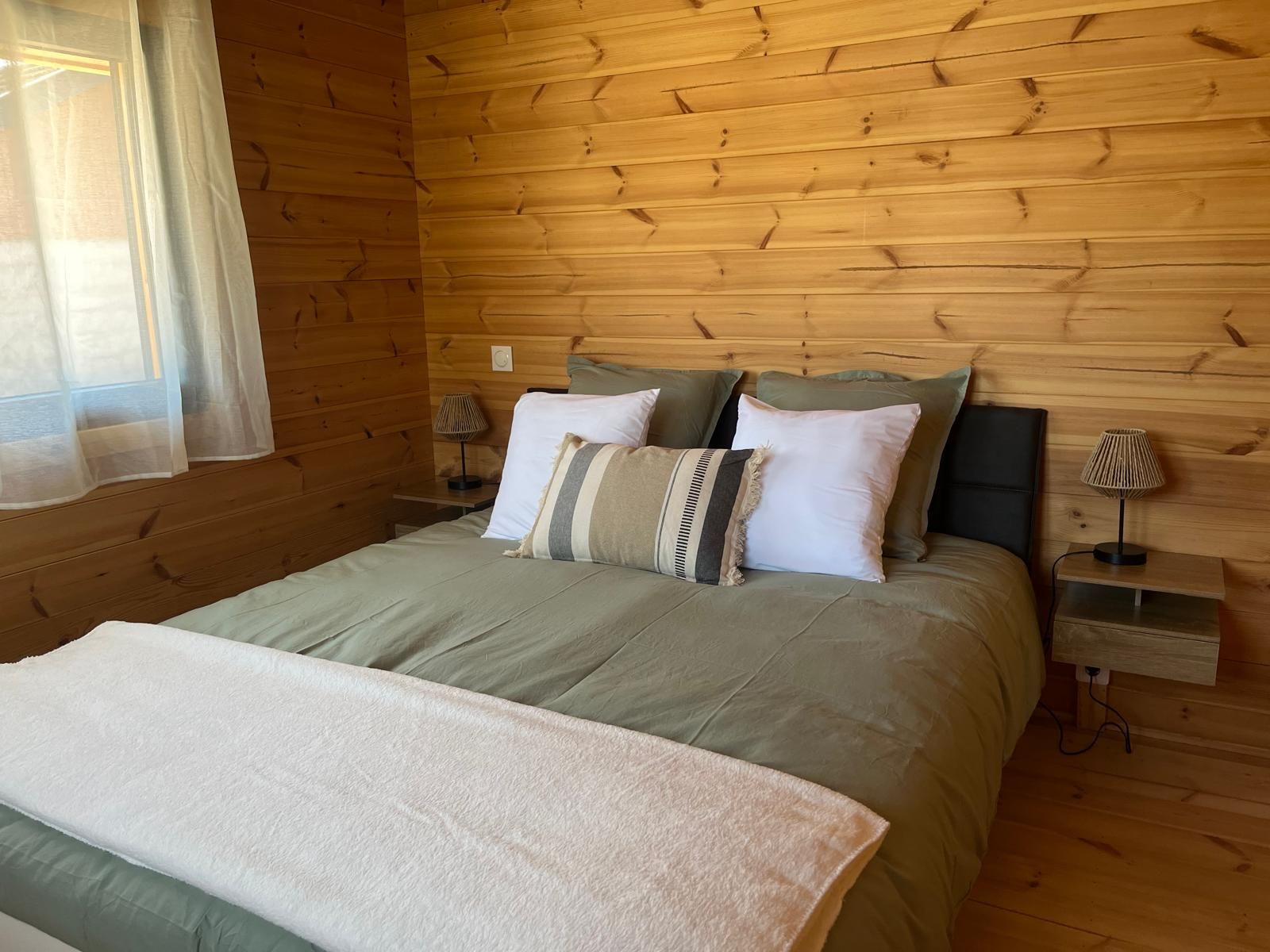 Une chambre à coucher aux murs lambrissés, avec un lit, deux tables de chevet et une fenêtre ornée de rideaux légers.