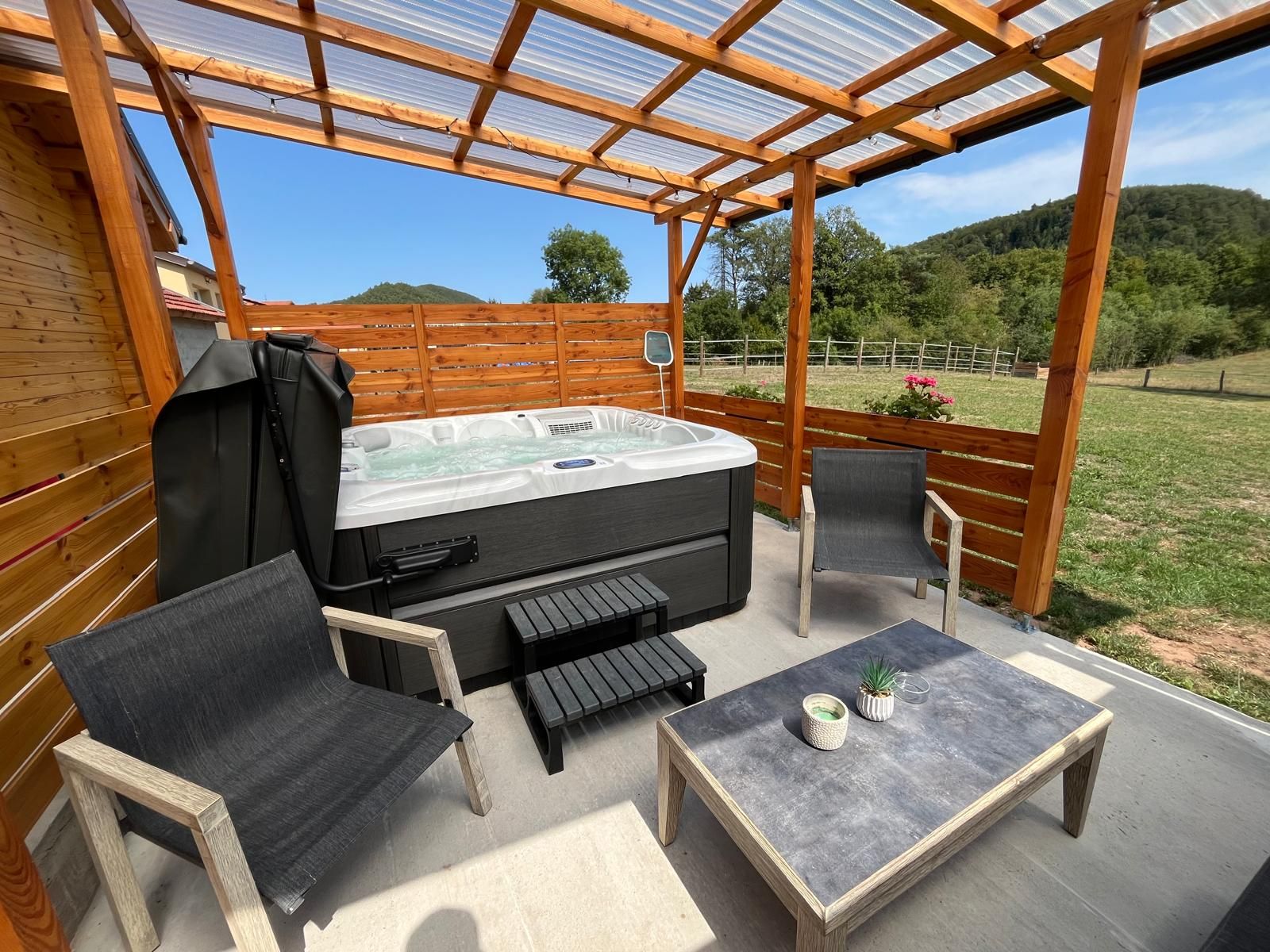 Een terras buiten met een hottub, twee stoelen, een tafel en een overdekte pergola.