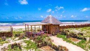 A beach hut with a thatched roof, surrounded by vegetation, and a sandy beach in the background.