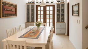 A dining area with a wooden table, white chairs, a vase with flowers, and a decorative rug. There are glass-fronted cabinets with dishes and a framed picture on the wall. - Maria Ines Bondesan (ubatuba)