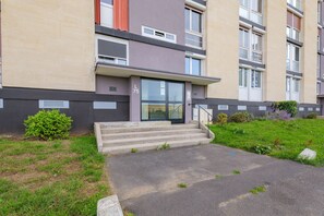 Un immeuble à plusieurs étages, doté d’un toit plat, d’un escalier menant à l’entrée et d’un petit jardin avec de la pelouse et des arbustes.
