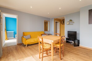 Un salon avec un canapé jaune, une table à manger en bois et des chaises, un téléviseur et une horloge au mur.