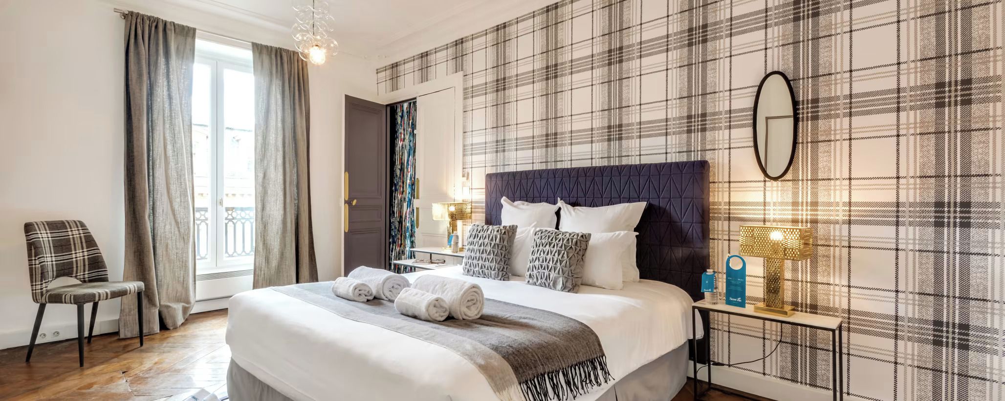 A hotel room with a bed, a chair, a bedside table with a lamp, and a window with curtains.
