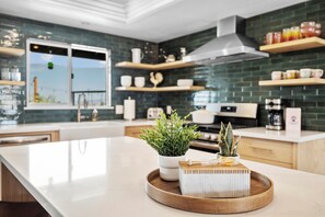 Eine moderne Küche mit einer großen Kochinsel, weißen Arbeitsplatten und einer grünen Spritzschutzwand. Darüber befinden sich Holzregale mit verschiedenen Küchenutensilien.