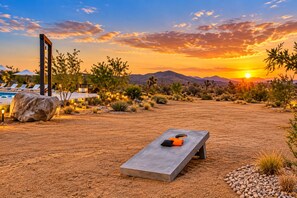 A desert landscape with a sunset, a pool area with lounge chairs, and a concrete bench with cushions. - Desert Shadow A 10ac Retreat w/Pool + Hot Tub+ Pickleball + Archery + More (Yucca Valley)