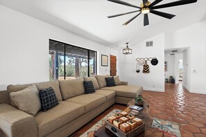A modern living room with a sectional sofa, a wooden coffee table, and a ceiling fan. - Desert Shadow A 10ac Retreat w/Pool + Hot Tub+ Pickleball + Archery + More (Yucca Valley)