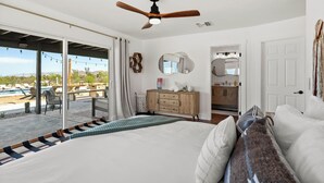 Slaapkamer met een groot bed, een plafondventilator, een houten dressoir en uitzicht op een zwembad en terras.