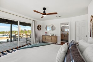 A bedroom with a large bed, a ceiling fan, a wooden dresser, and a view of a pool and outdoor area. - Desert Shadow A 10ac Retreat w/Pool + Hot Tub+ Pickleball + Archery + More (Yucca Valley)