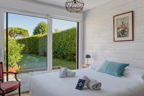 A bedroom with a large bed, a nightstand, a chair, and a view of a garden through a sliding door. - Mi Casa Blanca - piscine privée (La Teste-de-Buch)